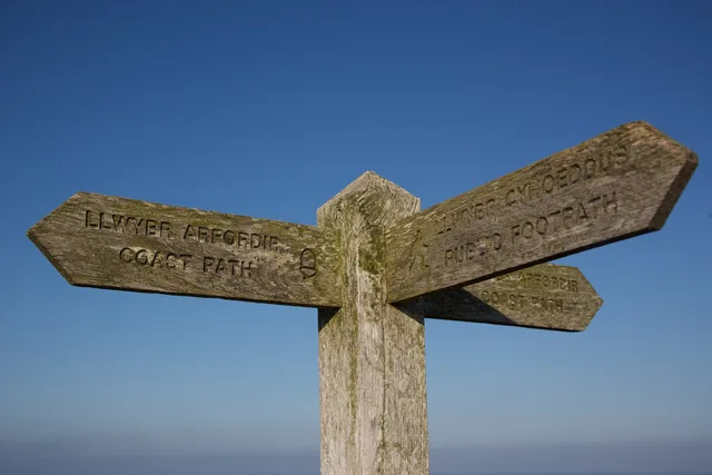 A rustic wooden signpost on the Welsh Coastal Path under a clear blue sky. Bilingual corporate communications in Wales: Welsh standards