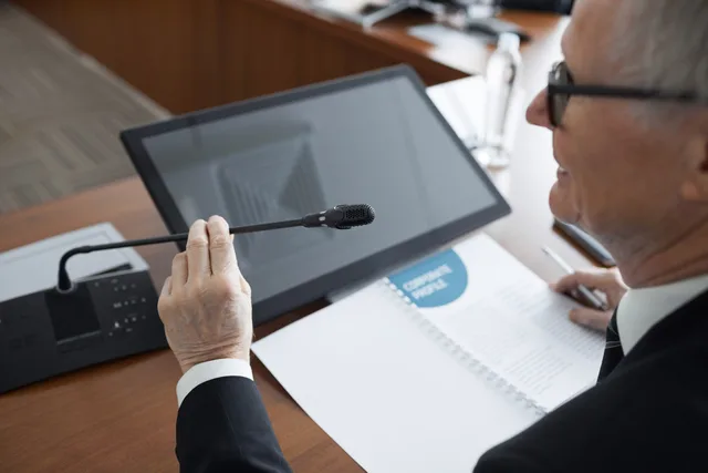 A senior businessman using a microphone during a conference meeting with documents and a monitor. Selecting corporate communications software only after the manual route is costed